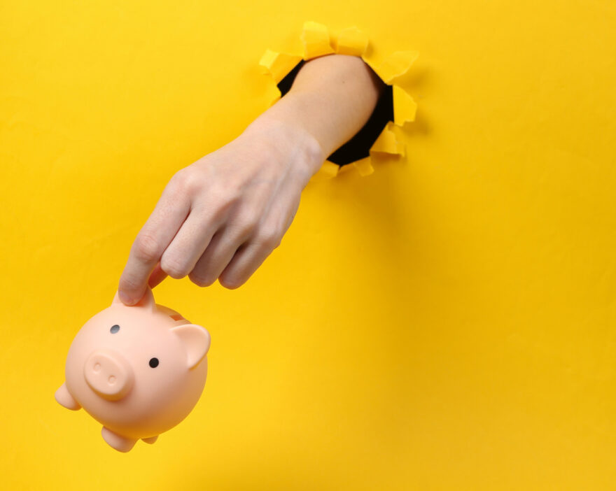 Female hand holds piggy bank through torn hole yellow paper.