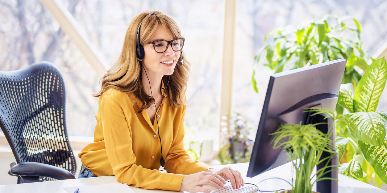 Mature businesswoman in headset speaking by conference call while looking at computer. Home office.