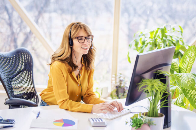 Mature businesswoman in headset speaking by conference call while looking at computer. Home office.