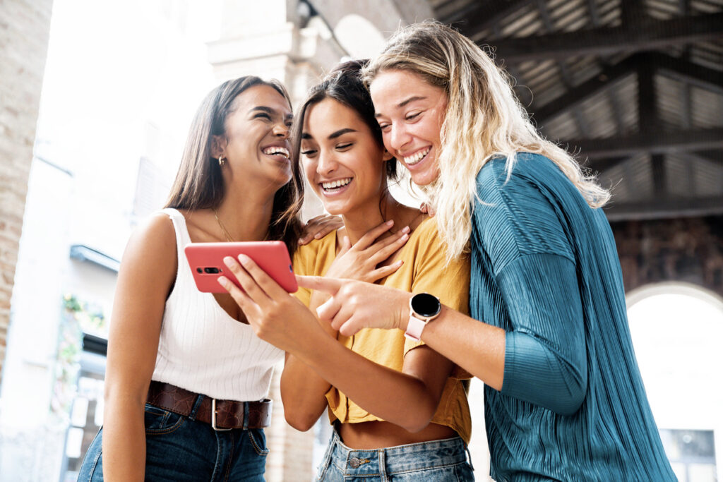 Three happy Gen Z friends watching a smart phone mobile outdoors - Millennials women using cellphone on city street - Technology, social, friendship and youth concept