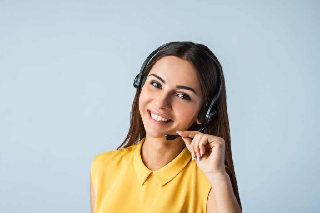 Photo of beautiful young call center operator for customer satisfaction standing near gray background. Woman with headphones looking at camera and smiling