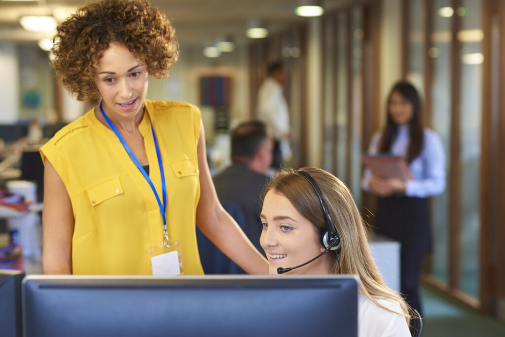 a young call center representative greets a caller in a large open plan office as her supervisor watches over her shoulder and helps her through the call.