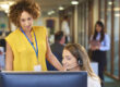 a young call center representative greets a caller in a large open plan office as her supervisor watches over her shoulder and helps her through the call.