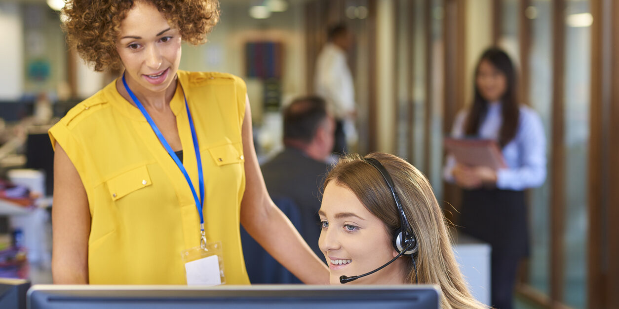 a young call center representative greets a caller in a large open plan office as her supervisor watches over her shoulder and helps her through the call.