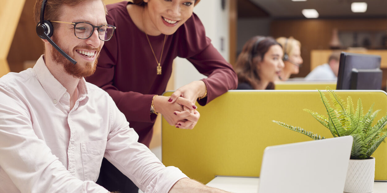 Businessman Wearing Headset Talking To Colleague In Busy Customer Services Centre Businessman Wearing Headset Talking To Colleague In Busy Customer Services Center for customer experience management
