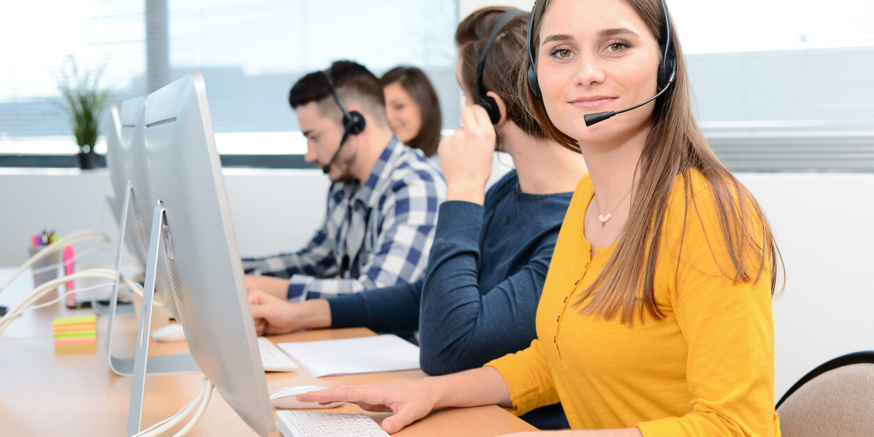 portrait of a beautiful and cheerful young woman telephone operator with headset working on desktop computer in row in a customer service call support helpline business center with teamworker in background portrait of a beautiful and cheerful young woman telephone operator with headset working on desktop computer in row in a customer service call support helpline business center with teamworker in background