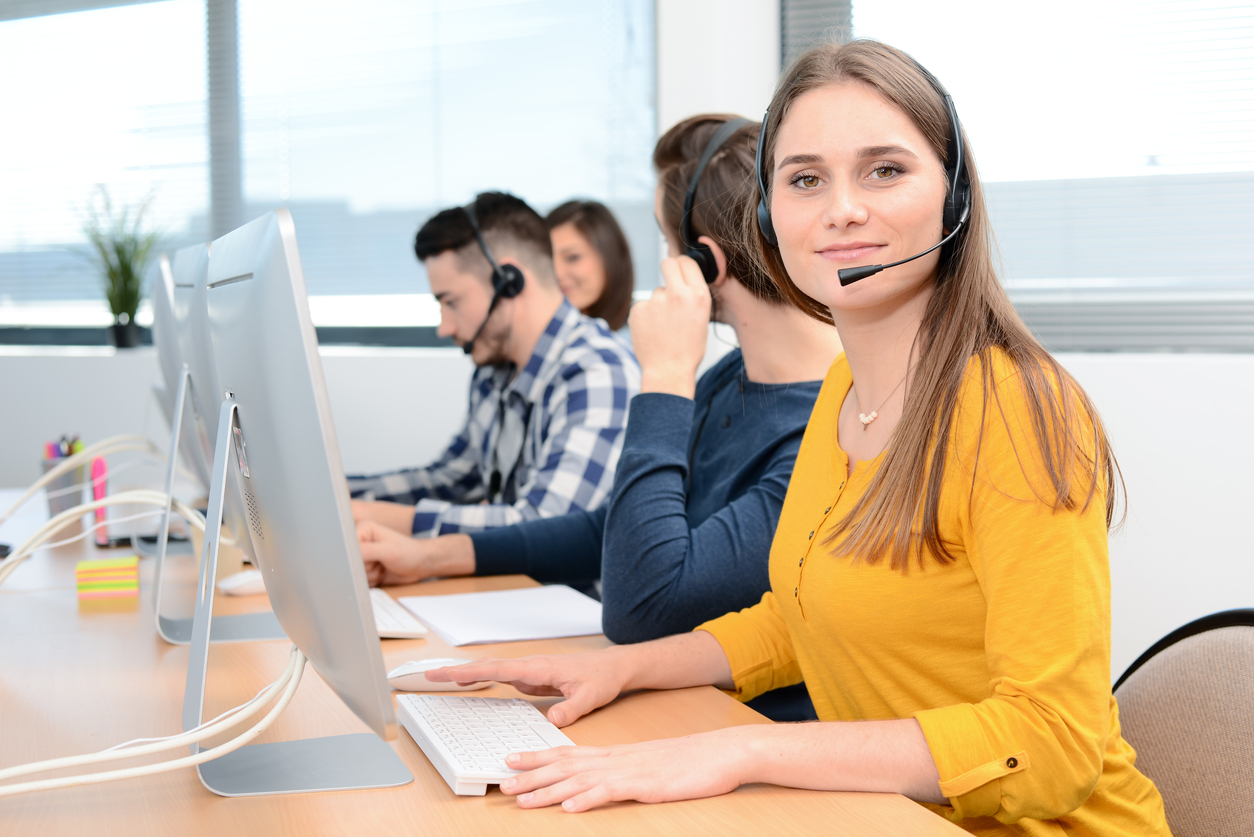 portrait of a beautiful and cheerful young woman telephone operator with headset working on desktop computer in row in a customer service call support helpline business center with teamworker in background