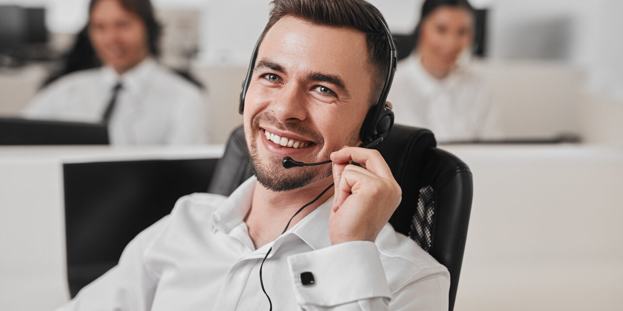 Positive operator with headset answering phone call Handsome smiling young male operator in headset consulting client during phone call while working in modern customer support service center