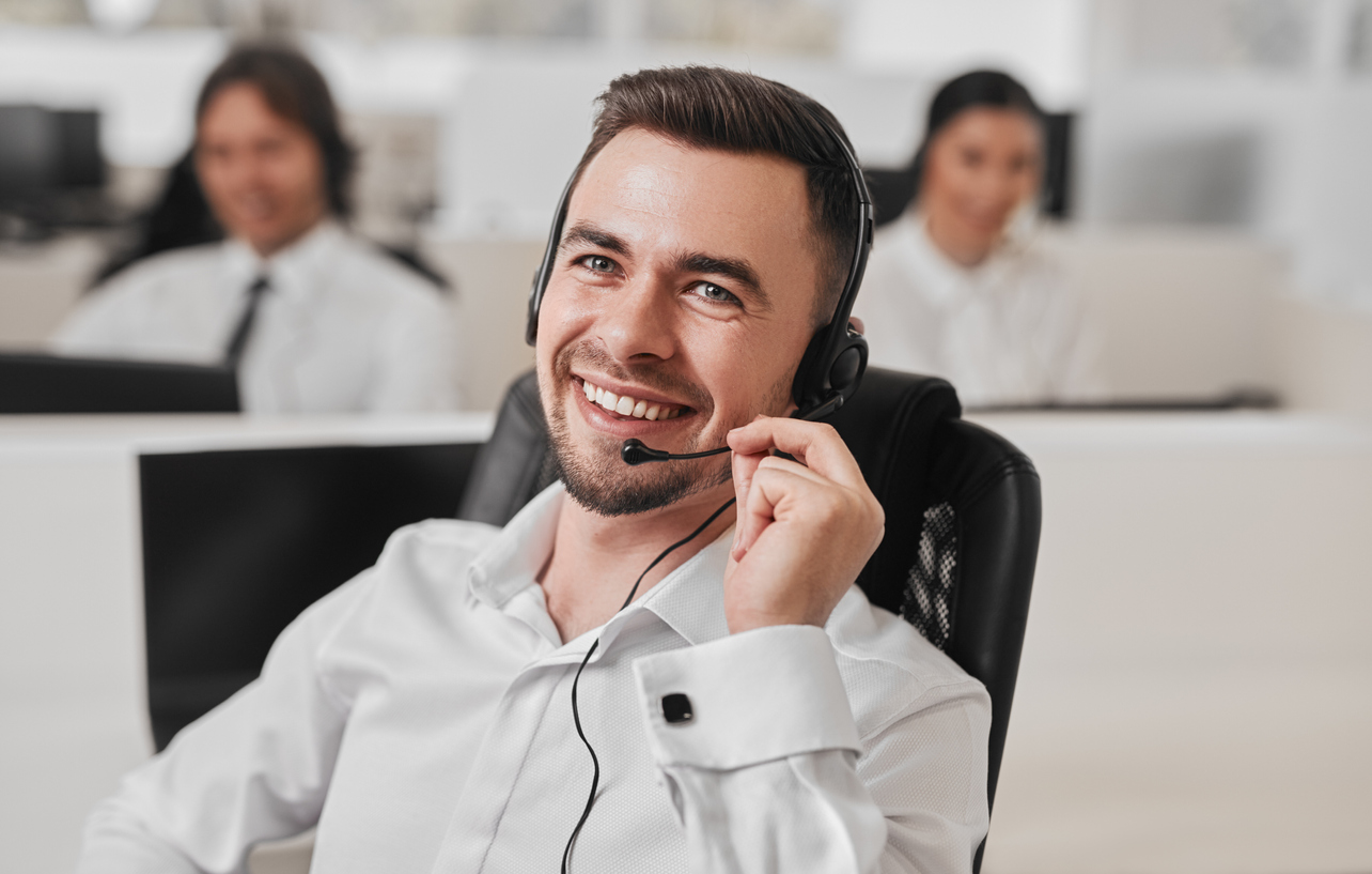 Handsome smiling young male operator in headset consulting client during phone call while working in modern customer support service center