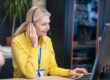 Blonde woman sitting in headphones at desk, typing, smiling