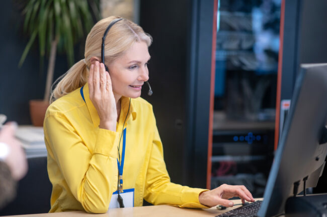 Blonde woman sitting in headphones at desk, typing, smiling
